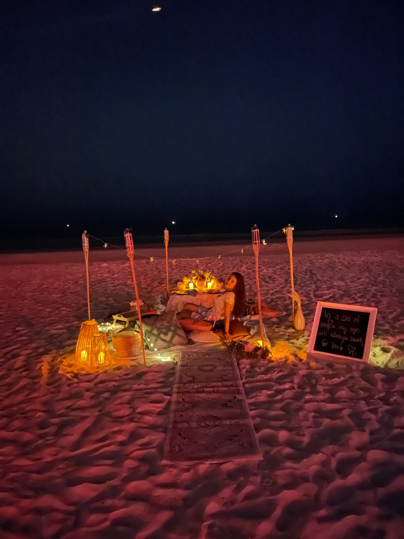 Noa under tiki torches at night with a chalkboard sign reading 'In a sea of people, my eyes will always search for you'